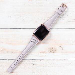 May include: A pale lilac Apple Watch strap with a rose gold case and buckle. The leather strap has a heart and arrow motif. The watch is shown on a light wood surface.