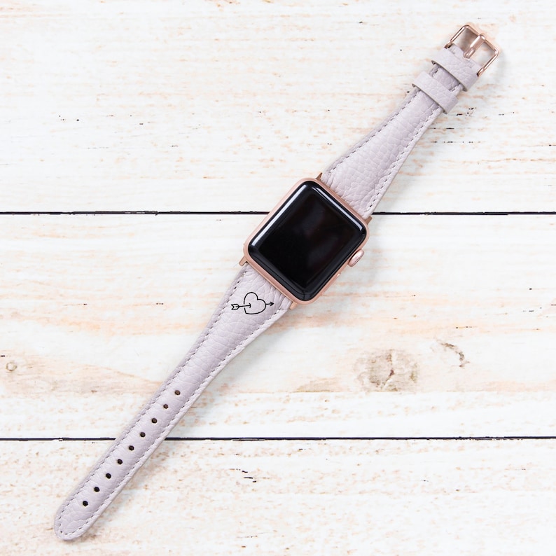 May include: A pale lilac Apple Watch strap with a rose gold case and buckle. The leather strap has a heart and arrow motif. The watch is shown on a light wood surface.