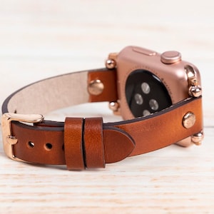 May include: A rose gold smartwatch with a brown leather strap. The strap has a gold buckle and is secured with gold rivets. The watch is displayed on a light wood surface.