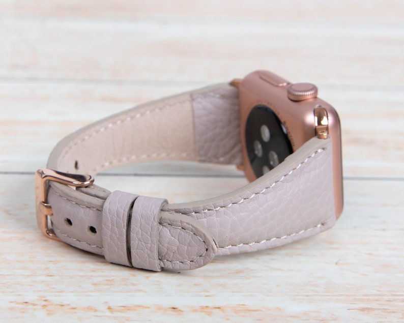 May include: A pale lilac leather watch strap with a rose gold buckle and watch case. The strap is stitched with white thread and has a textured surface. The watch case has a digital display.