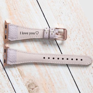 May include: A pale lilac leather watch strap with a rose gold buckle. The strap is engraved with the words "I love you" and a heart symbol. The watch strap is displayed on a light wood surface.