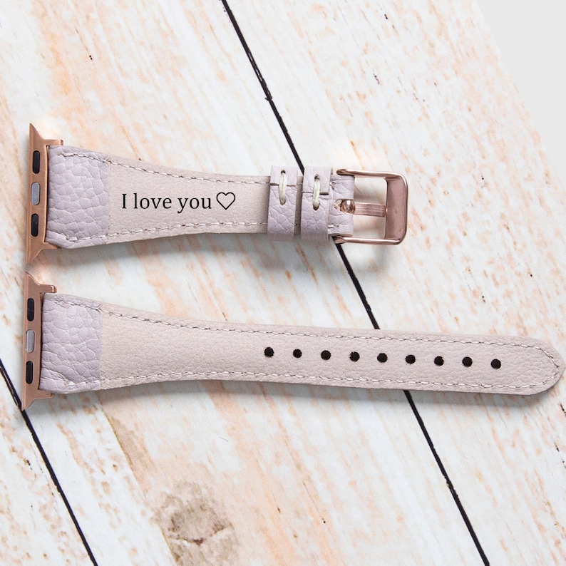 May include: A pale lilac leather watch strap with a rose gold buckle. The strap is engraved with the words "I love you" and a heart symbol. The watch strap is displayed on a light wood surface.