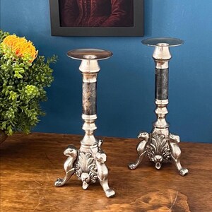 May include: Two silver candle holders with ornate bases and textured dark gray columns. The candle holders have a round top and are displayed on a wooden surface. A painting and a plant are in the background.