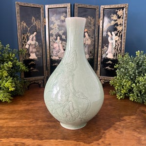 May include: A light green ceramic vase with a crackle glaze finish, featuring an etched design of a figure in traditional attire on the front. The vase has a bulbous body that tapers into a narrow neck and a smooth, rounded rim at the top.