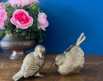 May include: Two gold-colored metal bird figurines. The birds are facing each other, with one bird perched on a branch and the other bird sitting on a nest.