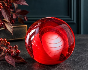 May include: A spherical glass paperweight featuring a deep red outer layer and a central white core with delicate, swirling horizontal lines encased within the transparent red glass, creating a layered, luminous visual effect.