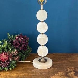 May include: A white and brass table lamp with a stacked orb design. The lamp features a round base and a series of four spherical, translucent orbs. The lamp has a brass neck and a gold-colored bulb fixture. The lamp is on a wooden surface.