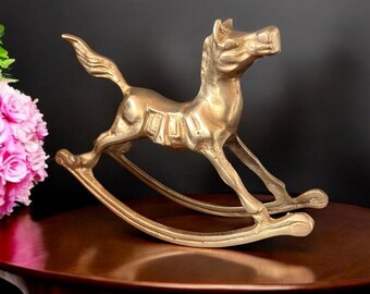 May include: A gold-colored metal rocking horse figurine. The horse is in a running pose with its head turned to the side. The rocking base is made of metal and has a smooth finish.