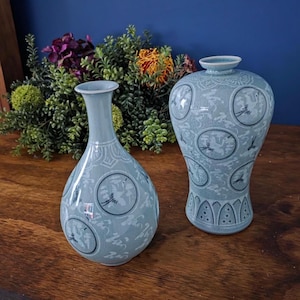 May include: Two light blue ceramic vases with intricate designs. The vases feature circular medallions with bird illustrations and detailed patterns. The vases are displayed on a wooden surface with greenery and flowers in the background.