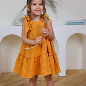 Könnte beinhalten: Ein senfgelbes ärmelloses Kleid mit quadratischem Ausschnitt, zum Binden an den Schultern und einem gestuften Rock mit gerüschter Naht. Das Kleid besteht aus strukturiertem Stoff und wird mit braunen Schnallensandalen und einer gelben Blume getragen.