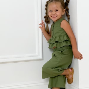 Puede incluir: Una niña pequeña con un conjunto de dos piezas verde oliva, con un top con volantes y pantalones de pernera ancha. La niña también lleva sandalias marrones. El conjunto es de tela suave.