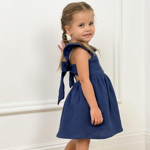 May include: A young girl wearing a navy blue dress with a bow detail on the back. The dress has a flared skirt and is paired with black flats with pearl accents. The girl is smiling and standing in front of a white wall.