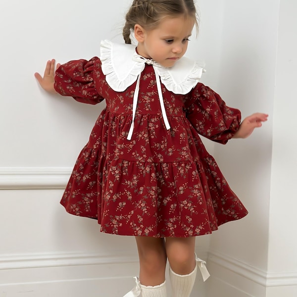 May include: A red floral print dress with a white ruffled collar and long sleeves. The dress is paired with white knee-high socks and black Mary Jane shoes. The dress has a flared skirt.