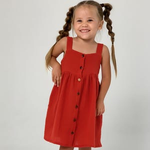 Peut inclure: Une jeune fille porte une robe rouge boutonnée avec un col carré et de larges bretelles. Elle porte des sandales marron. La fille sourit, ses cheveux sont tressés en deux nattes.
