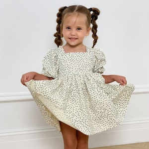 Puede incluir: Una niña pequeña con un vestido crema con cuello cuadrado y mangas cortas abullonadas. El vestido está cubierto de pequeños estampados florales azules. La niña sonríe y levanta la falda del vestido, revelando sus piernas desnudas.