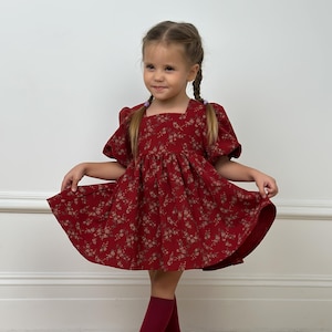 Puede incluir: Un vestido rojo oscuro con escote cuadrado, mangas cortas abullonadas y un delicado estampado floral blanco. El vestido tiene la cintura fruncida y una falda acampanada. El conjunto se completa con un par de calcetines altos de color rojo oscuro.