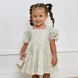 以下が含まれることがあります： クリーム色のドレスを着た少女。スクエアネックと短いパフスリーブが特徴です。ドレスには繊細な青い花柄が施されています。少女は笑顔で、髪は二つのおさげに結ばれています。