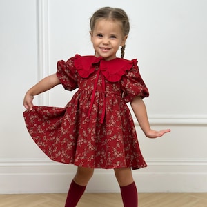 May include: A red floral print dress with a ruffled collar and matching knee-high socks. The dress features short, puffed sleeves and a tie at the neckline. The girl is smiling and has her arms outstretched.