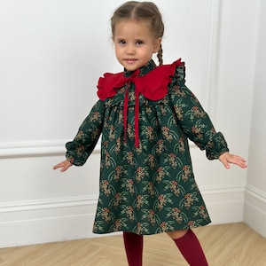 May include: A green dress with a floral pattern and a red ruffled collar. The dress has long sleeves and a red ribbon tie. The outfit is completed with burgundy tights and black flats with pearl accents. The dress is for a child.