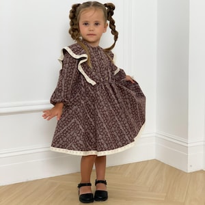 Könnte beinhalten: Ein junges Mädchen in einem braunen Kleid mit floralem Muster, Rüschenkragen und weißer Spitzenborte. Sie trägt schwarze Mary Jane Schuhe. Ihr Haar ist zu zwei geflochtenen Zöpfen geflochten. Das Kleid ist knielang.