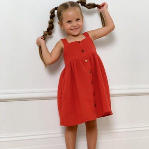 May include: A young child in a red dress with a square neckline and button-down front, paired with brown sandals. The child smiles, holding up their braided hair. The background is a plain white wall. The dress is a vibrant shade of red.