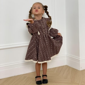 Może przedstawiać: Brązowa sukienka w kwiaty z białym koronkowym wykończeniem. Sukienka ma długie rękawy i okrągły dekolt. Dziecko ma na sobie czarne buty Mary Jane. Tło to gładka biała ściana.