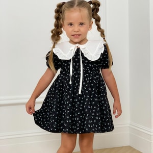 May include: A black floral print dress with a white Peter Pan collar and short sleeves. The dress features a flared skirt and is worn with black Mary Jane shoes. The girl has blonde hair styled in pigtails.