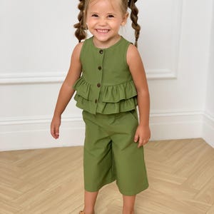 May include: A young child wearing a two-piece olive green outfit. The top is a sleeveless button-down with a ruffled hem. The matching culottes are knee-length. The child is also wearing brown sandals.