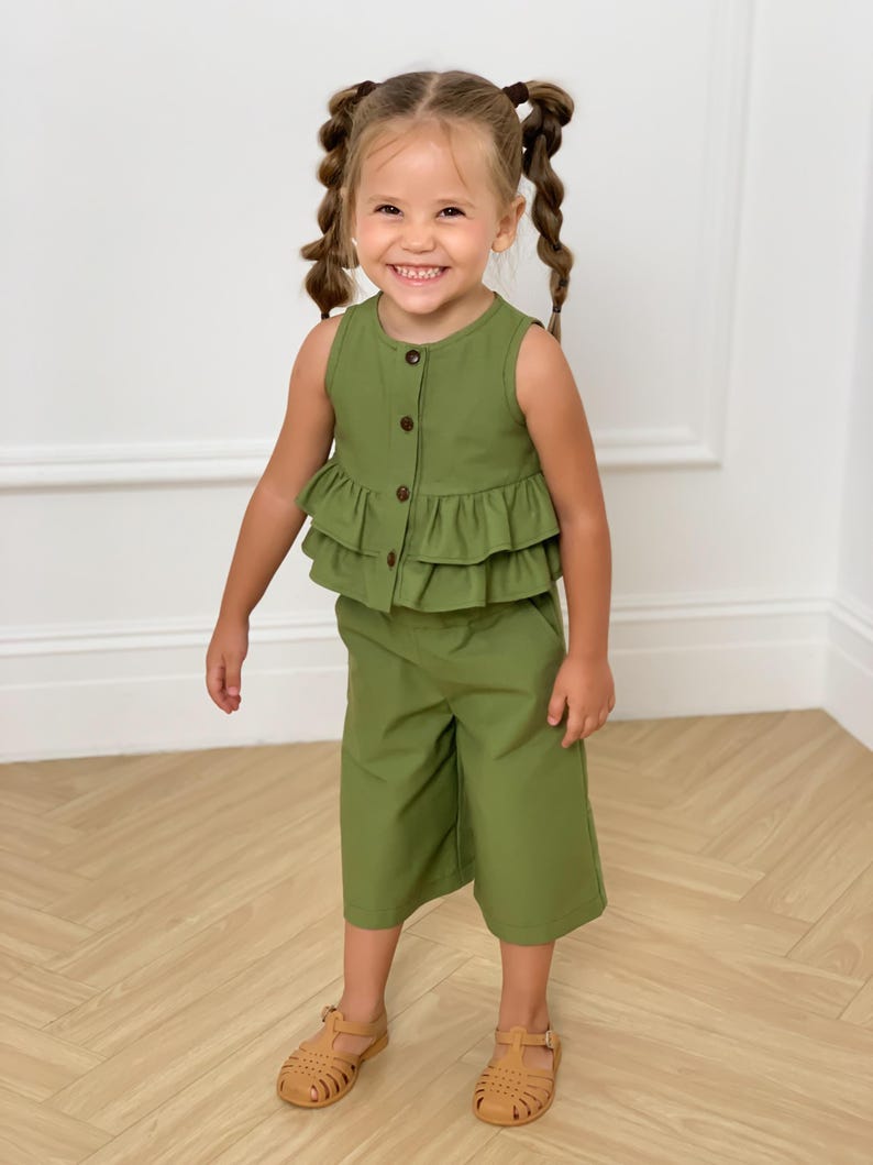 May include: A young child wearing a two-piece olive green outfit. The top is a sleeveless button-down with a ruffled hem. The matching culottes are knee-length. The child is also wearing brown sandals.