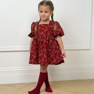 Peut inclure: Une jeune fille porte une robe rouge à motifs floraux et des chaussettes hautes assorties. La robe a des manches courtes bouffantes et une encolure carrée. Les chaussettes sont d'un rouge profond.
