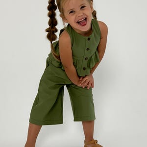 May include: A young child wearing a green sleeveless top with buttons and matching cropped pants. The outfit is paired with brown sandals. The child is smiling and has braided pigtails.