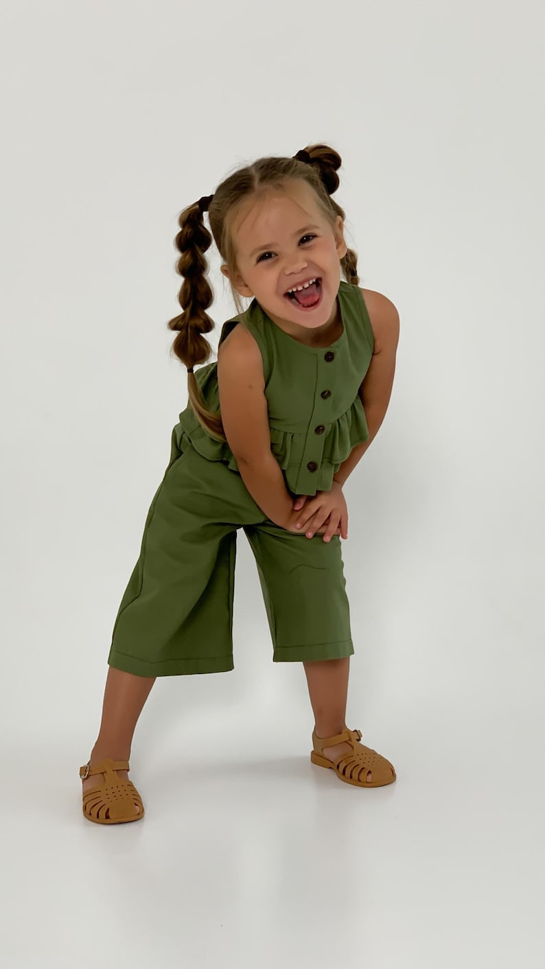 May include: A young child wearing a green sleeveless top with buttons and matching cropped pants. The outfit is paired with brown sandals. The child is smiling and has braided pigtails.