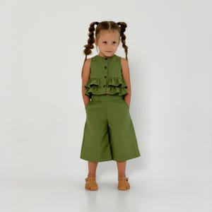 May include: A young girl wearing a two-piece olive green outfit. The top is a sleeveless button-down with layered ruffles. The matching pants are wide-legged culottes. She is wearing brown sandals.