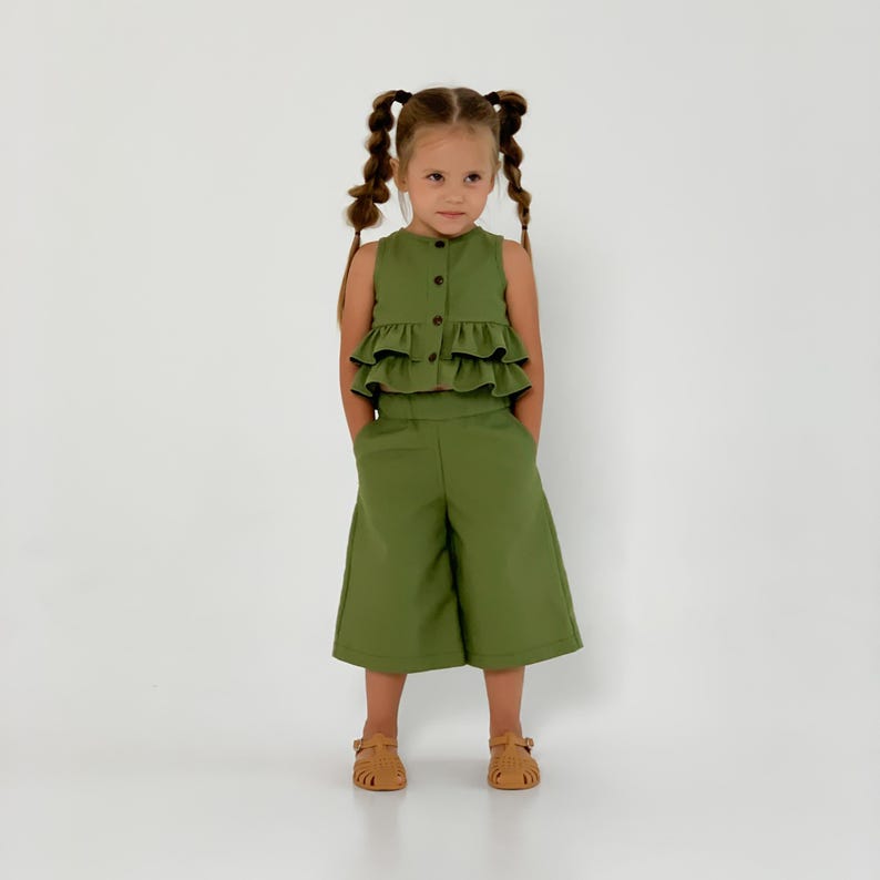 May include: A young girl wearing a two-piece olive green outfit. The top is a sleeveless button-down with layered ruffles. The matching pants are wide-legged culottes. She is wearing brown sandals.