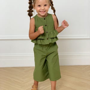 May include: A young child is wearing a two-piece olive green outfit. The top is a sleeveless button-down with a ruffled hem. The matching pants are cropped. The child is also wearing brown sandals.