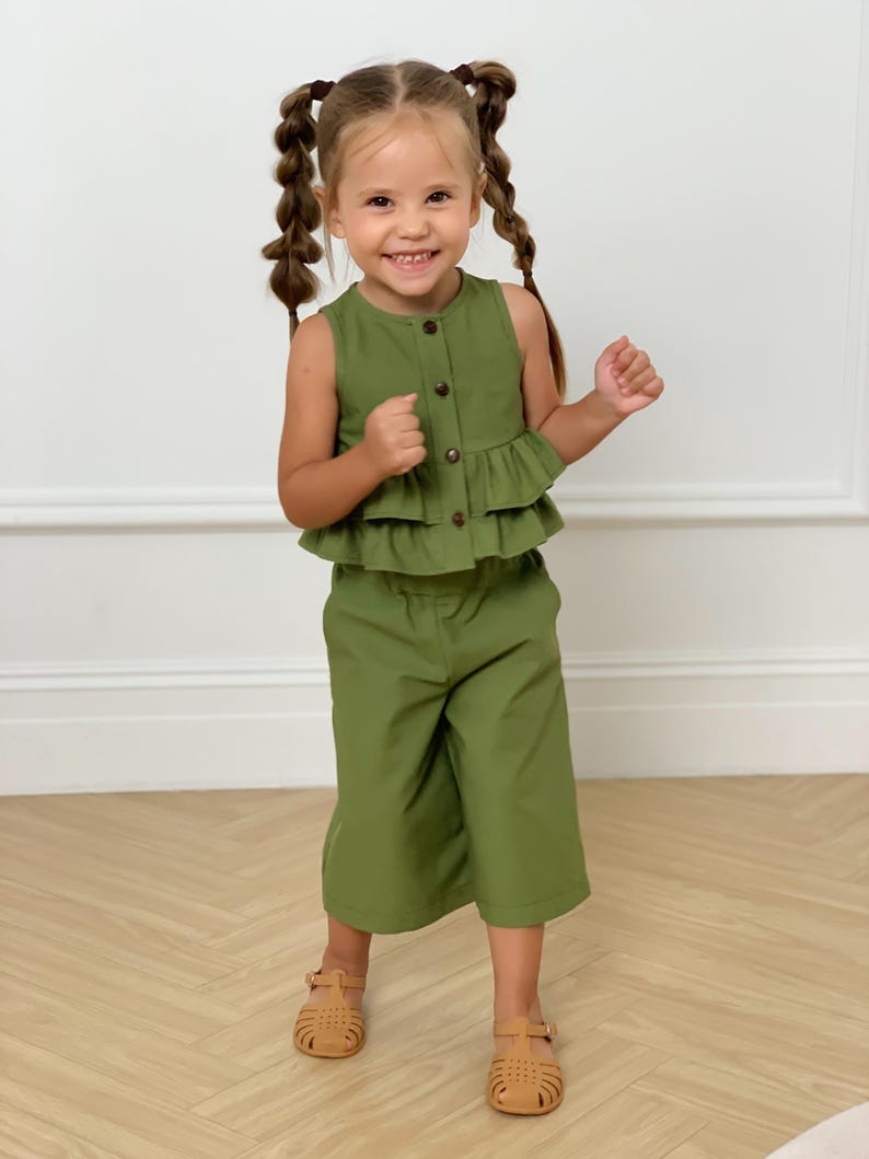 May include: A young child is wearing a two-piece olive green outfit. The top is a sleeveless button-down with a ruffled hem. The matching pants are cropped. The child is also wearing brown sandals.
