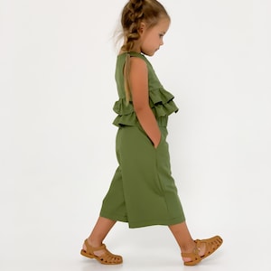 May include: A young child wearing an olive green outfit with a ruffled top and cropped pants. The outfit has pockets. The child is wearing brown sandals. The background is white.