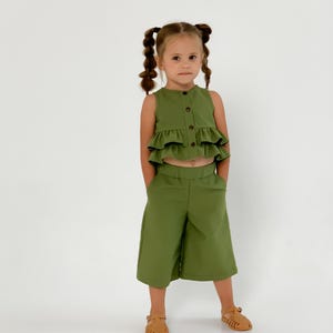 May include: A green two-piece outfit for a child. The top is a sleeveless, button-down shirt with ruffled layers. The matching pants are cropped culottes. The child is wearing brown sandals.