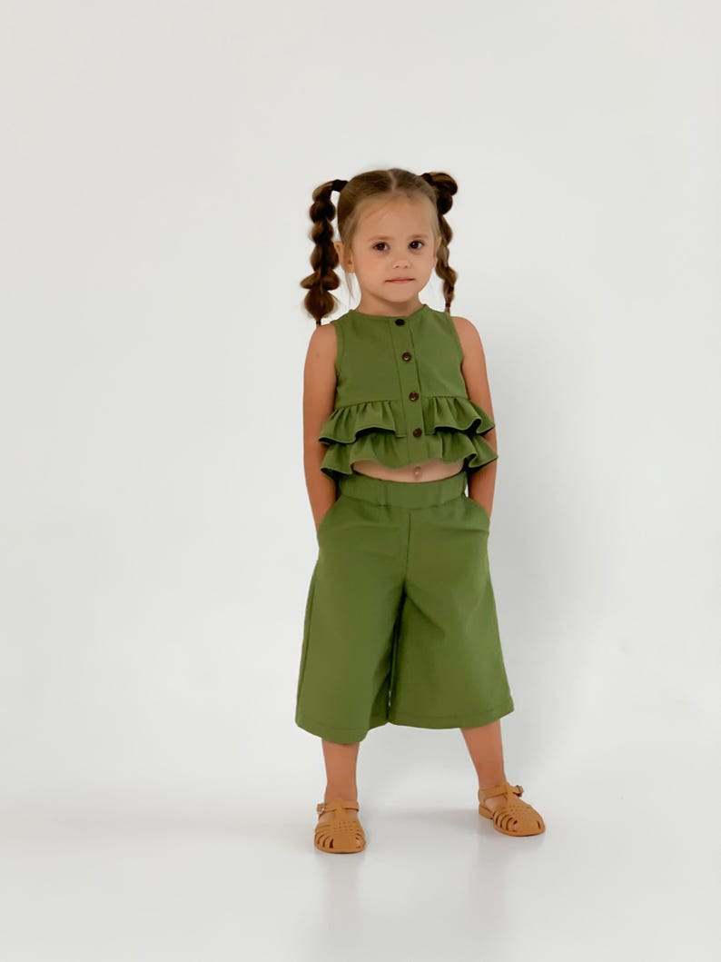 May include: A green two-piece outfit for a child. The top is a sleeveless, button-down shirt with ruffled layers. The matching pants are cropped culottes. The child is wearing brown sandals.
