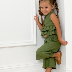May include: A young child wearing a two-piece olive green outfit with a ruffled top and cropped pants. The child is also wearing brown sandals. The outfit is a solid color and appears to be made of a soft fabric.