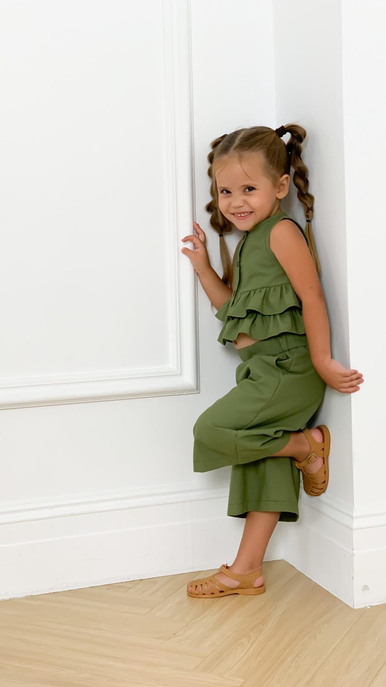 May include: A young child wearing a two-piece olive green outfit with a ruffled top and cropped pants. The child is also wearing brown sandals. The outfit is a solid color and appears to be made of a soft fabric.