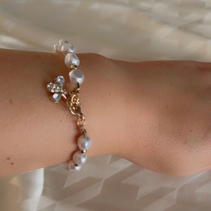 Op de afbeelding: Een witte parel armband met een gouden ketting en een kleine vlinder bedel met strass.
