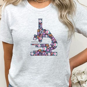 May include: A gray t-shirt with a floral patterned microscope design. The microscope is outlined in dark blue and the text "Hematology" is printed on the microscope.