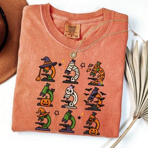 May include: A coral-colored t-shirt featuring a Halloween-themed design. The design includes nine illustrations of microscopes decorated with witch hats, bats, pumpkins, and candy. A gold necklace with a pendant is draped over the shirt.