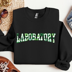 Può includere: Felpa nera con la scritta "LABORATORY" in bianco, riempita di trifogli verdi. Un capo comodo, ideale per le celebrazioni di San Patrizio.
