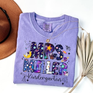May include: A light purple Comfort Colors t-shirt with the words "MRS MILLER Kindergarten" in a space-themed design. The design includes planets, stars, rockets, and an astronaut. A gold necklace is draped across the shirt.