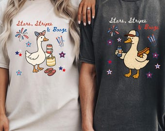 May include: Two t-shirts with a patriotic theme. One is cream-colored, the other is dark gray. Both feature a cartoon duck with a beer and the text "Stars, Stripes & Booze", along with fireworks and star graphics.