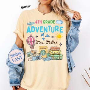 May include: A butter yellow t-shirt featuring a graphic print with the text 4TH GRADE ADVENTURE and Mrs Miller. The design includes a hot air balloon, a camera, a passport, stacked suitcases, a sun, clouds, an airplane, and birds.