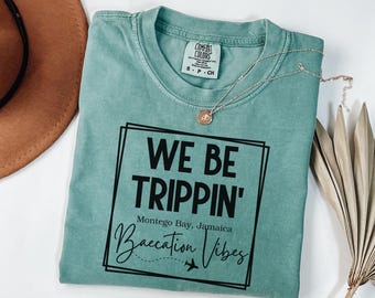 Op de afbeelding: Een tealblauw T-shirt met de tekst "We Be Trippin'" in zwart. De tekst staat in een zwarte rechthoek met een witte rand. Onder de tekst staat "Montego Bay, Jamaica" en "Baecation Vibes" in een cursief schrift met een klein vliegtuigicoon.