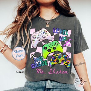 Puede incluir: Una camiseta gris oscuro con tres coloridos mandos de videojuegos. La camiseta tiene las palabras "LEVEL UP" y "2ND GRADE Ms. Sharon" en una fuente divertida. Un patr&oacute;n de cuadros rosas y negros est&aacute; en el fondo.